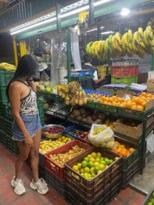 Mujer seleccionando frutas frescas en un mercado local con bananas colgadas y variedad de cítricos, uvas y cocos en cajas de colores."